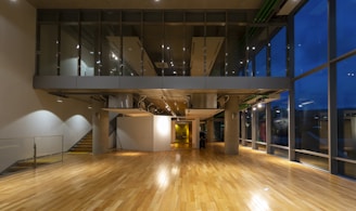 A modern interior space features wooden flooring and large glass windows that extend from floor to ceiling, allowing a view of the exterior nighttime scene. Suspended overhead, there is a mezzanine level with a glass facade, and visible ductwork adds an industrial touch. The area is well-lit with overhead lighting, and the space appears spacious and open.