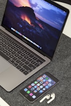A MacBook Pro displaying a vibrant lava-themed wallpaper is placed on a grey textured surface. In front of the laptop, an iPhone displays various app icons on its home screen. Beside the iPhone, a pair of white AirPods are resting on the surface.