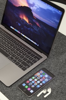 A MacBook Pro displaying a vibrant lava-themed wallpaper is placed on a grey textured surface. In front of the laptop, an iPhone displays various app icons on its home screen. Beside the iPhone, a pair of white AirPods are resting on the surface.