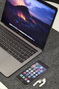 A MacBook Pro displaying a vibrant lava-themed wallpaper is placed on a grey textured surface. In front of the laptop, an iPhone displays various app icons on its home screen. Beside the iPhone, a pair of white AirPods are resting on the surface.