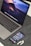 A MacBook Pro displaying a vibrant lava-themed wallpaper is placed on a grey textured surface. In front of the laptop, an iPhone displays various app icons on its home screen. Beside the iPhone, a pair of white AirPods are resting on the surface.