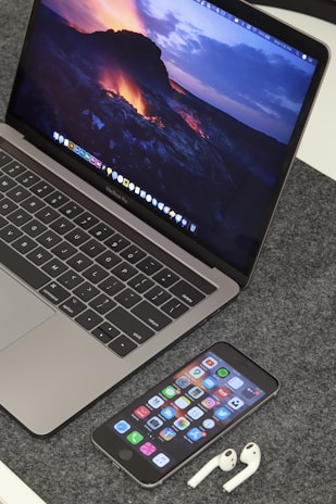 A MacBook Pro displaying a vibrant lava-themed wallpaper is placed on a grey textured surface. In front of the laptop, an iPhone displays various app icons on its home screen. Beside the iPhone, a pair of white AirPods are resting on the surface.