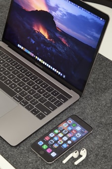 A MacBook Pro displaying a vibrant lava-themed wallpaper is placed on a grey textured surface. In front of the laptop, an iPhone displays various app icons on its home screen. Beside the iPhone, a pair of white AirPods are resting on the surface.