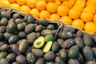 A display of fresh produce with a foreground of dark avocados, some whole and one cut in half revealing green interior, and a background of bright orange tangerines stacked in a neat arrangement.