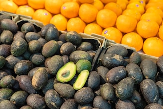 A display of fresh produce with a foreground of dark avocados, some whole and one cut in half revealing green interior, and a background of bright orange tangerines stacked in a neat arrangement.