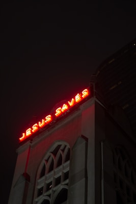 A building with an illuminated red neon sign reading 'JESUS SAVES' at the top, against a dark night sky. The structure has multiple large arched windows, giving it a classical architectural appearance.