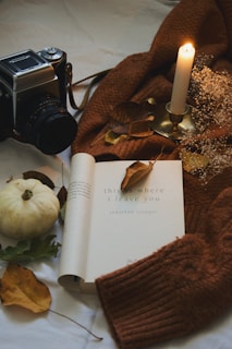 A cozy setup including an open book, a vintage camera, a lit candle in a holder, and an arrangement of autumn leaves. A small white pumpkin and a brown knit sweater add to the warm, inviting atmosphere.