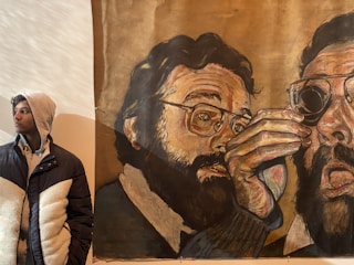 A young person wearing a hooded jacket stands beside a large, expressive mural. The mural features a detailed depiction of a bearded man with eyeglasses, partly obscured by his own hand in a dramatic pose.