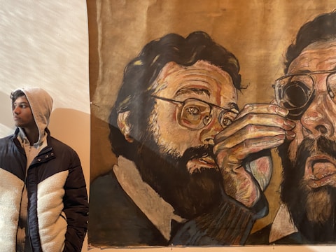 A young person wearing a hooded jacket stands beside a large, expressive mural. The mural features a detailed depiction of a bearded man with eyeglasses, partly obscured by his own hand in a dramatic pose.