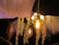 An elegantly decorated ceiling features a hanging light fixture with exposed filament bulbs, creating a warm and cozy ambiance. Cascading floral garlands and what appear to be crystal bead strands hang from the ceiling, enhancing the decorative nature of the setting. The warm glow from the bulbs casts gentle light on the decor, adding to the atmosphere. A person is partially visible in the foreground, with attention focused on the decor.