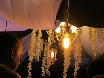 An elegantly decorated ceiling features a hanging light fixture with exposed filament bulbs, creating a warm and cozy ambiance. Cascading floral garlands and what appear to be crystal bead strands hang from the ceiling, enhancing the decorative nature of the setting. The warm glow from the bulbs casts gentle light on the decor, adding to the atmosphere. A person is partially visible in the foreground, with attention focused on the decor.