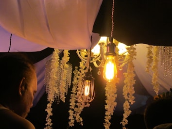 An elegantly decorated ceiling features a hanging light fixture with exposed filament bulbs, creating a warm and cozy ambiance. Cascading floral garlands and what appear to be crystal bead strands hang from the ceiling, enhancing the decorative nature of the setting. The warm glow from the bulbs casts gentle light on the decor, adding to the atmosphere. A person is partially visible in the foreground, with attention focused on the decor.