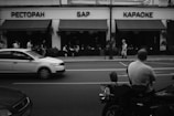 A monochrome photograph of a street scene with a restaurant, bar, and karaoke building in the background. Several people are seen sitting at tables under the awning, while pedestrians walk along the sidewalk. In the foreground, a man is sitting on a motorcycle facing the street, and a car is driving past.