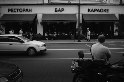 A monochrome photograph of a street scene with a restaurant, bar, and karaoke building in the background. Several people are seen sitting at tables under the awning, while pedestrians walk along the sidewalk. In the foreground, a man is sitting on a motorcycle facing the street, and a car is driving past.