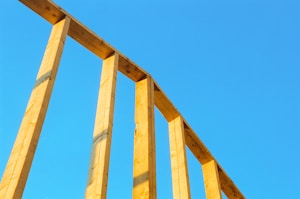Wooden beams form a simple frame against a clear blue sky. The structure is partially constructed, with vertical and horizontal elements creating a geometric pattern.