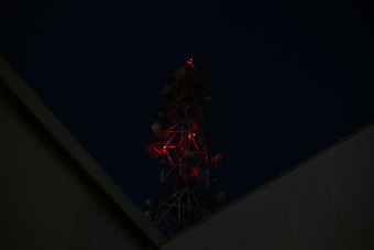 A dark night scene featuring a tall communication tower with red lights that glow in the darkness. The silhouette of the tower is visible against the night sky, surrounded by building structures.