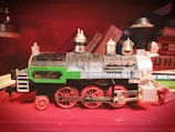 A vintage toy train model with intricate detailing and the label 'Denver Express' on its side. The train is primarily black with green and silver accents, and red wheels. It is placed against a background of colorful stacked books, adding a retro and cozy atmosphere.
