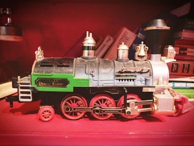 A vintage toy train model with intricate detailing and the label 'Denver Express' on its side. The train is primarily black with green and silver accents, and red wheels. It is placed against a background of colorful stacked books, adding a retro and cozy atmosphere.