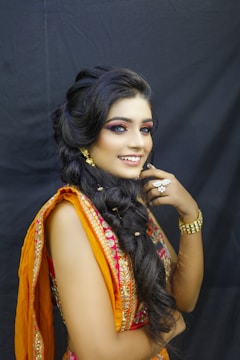 A woman with dark, styled hair adorned with gold ornaments is smiling while wearing an ornate traditional dress. The outfit features intricate embroidery in orange and pink, complementing her eye makeup. She is accessorized with gold jewelry, including earrings, a bracelet, and a ring.