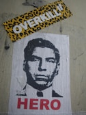 A stencil-style poster of a man's face is displayed on a wall. Below the image, the word 'HERO' is written in bold red letters. Above the poster, there is a sticker with a leopard print background and the word 'OVERKILL' in bold white letters.