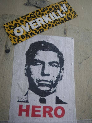 A stencil-style poster of a man's face is displayed on a wall. Below the image, the word 'HERO' is written in bold red letters. Above the poster, there is a sticker with a leopard print background and the word 'OVERKILL' in bold white letters.