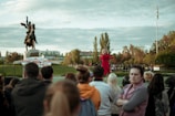 A person in a red outfit is speaking to an audience outdoors. The audience is facing the speaker and consists of people of various ages and genders. In the background, there is a statue of a person on horseback, trees, and a flagpole with a flag. The setting appears to be a park or open area with cloudy skies above.
