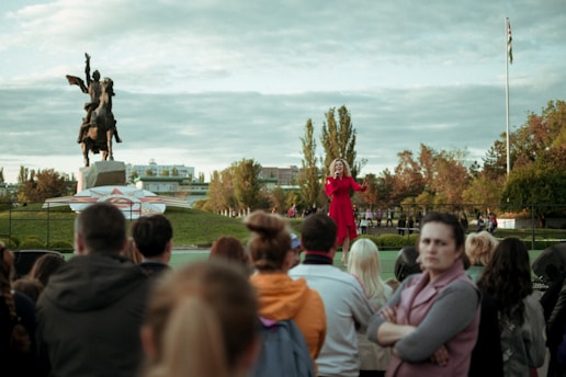 A person in a red outfit is speaking to an audience outdoors. The audience is facing the speaker and consists of people of various ages and genders. In the background, there is a statue of a person on horseback, trees, and a flagpole with a flag. The setting appears to be a park or open area with cloudy skies above.