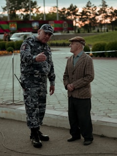 A man in camouflage uniform, appearing to be a police officer, is engaged in conversation with an elderly man who is wearing a plaid blazer and cap. They are standing on a paved area with greenery and flag decorations in the background, suggesting an outdoor public space.