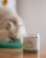 A fluffy cat is eating from a bowl in the background, while in the foreground there is a jar labeled Botaneo with cat treats inside. The setting appears to be indoors with soft lighting.