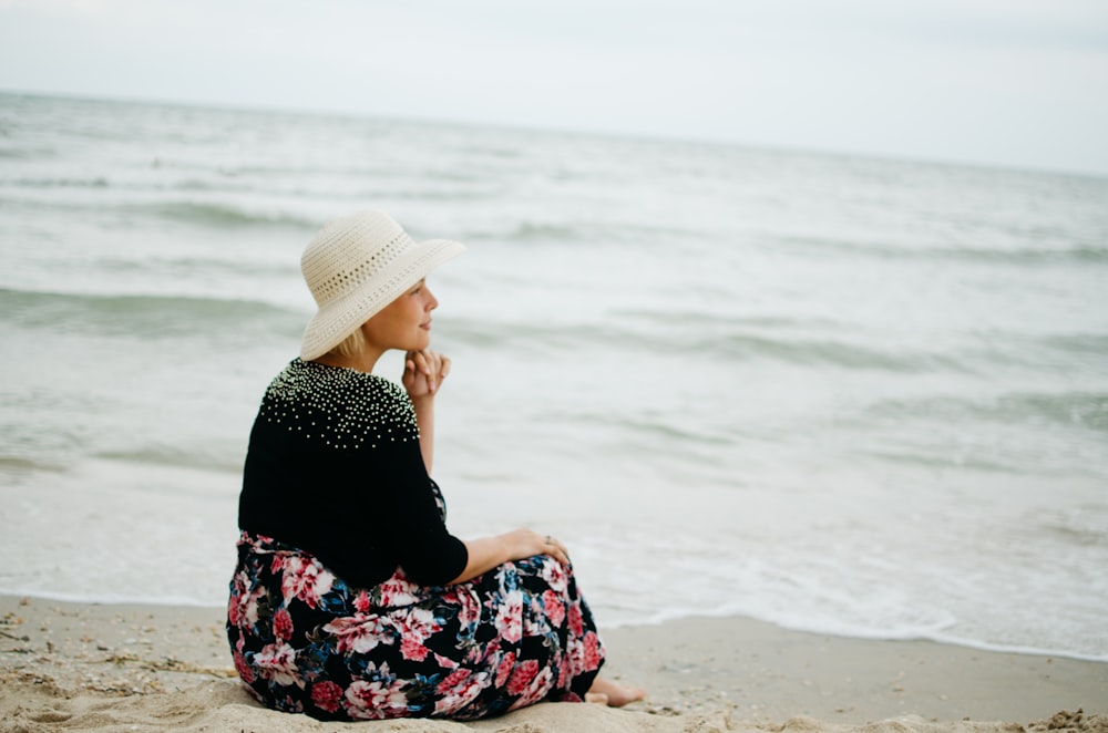 woman in black and red floral dress wearing white fedora hat sitting on seashore during daytime