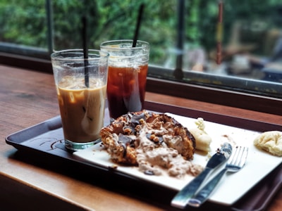 Two glasses with iced coffee drinks are placed on a tray next to a partially eaten dessert. The scene is set indoors, by a window overlooking greenery, creating a relaxed café atmosphere. The drinks have black straws, and one is a light brown shade while the other is darker. A fork and knife are on the tray as well.
