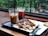 Two glasses with iced coffee drinks are placed on a tray next to a partially eaten dessert. The scene is set indoors, by a window overlooking greenery, creating a relaxed café atmosphere. The drinks have black straws, and one is a light brown shade while the other is darker. A fork and knife are on the tray as well.
