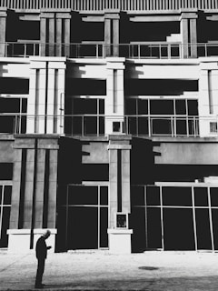 A person wearing a hard hat is standing in front of a modern building facade with strong geometric architectural lines. The building features tall columns and large glass windows, creating an urban and industrial aesthetic. The image is in black and white, enhancing the contrast between light and shadow.
