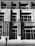 A person wearing a hard hat is standing in front of a modern building facade with strong geometric architectural lines. The building features tall columns and large glass windows, creating an urban and industrial aesthetic. The image is in black and white, enhancing the contrast between light and shadow.
