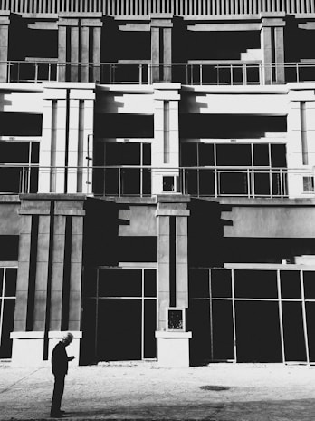 A person wearing a hard hat is standing in front of a modern building facade with strong geometric architectural lines. The building features tall columns and large glass windows, creating an urban and industrial aesthetic. The image is in black and white, enhancing the contrast between light and shadow.