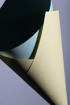 A close-up of a paper sculpture composed of sheets forming layered curves. Each sheet displays subtle shades of green and blue with soft light creating delicate shadows.