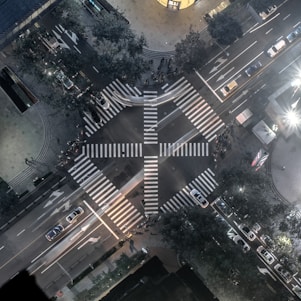 An aerial view of an urban intersection featuring a large, cross-shaped pedestrian crosswalk. The streets are lined with various vehicles, including cars and buses. Trees and buildings border the sidewalks, with some illuminated signage and streetlights visible. The overall scene is bustling yet orderly, with both foot and vehicle traffic.