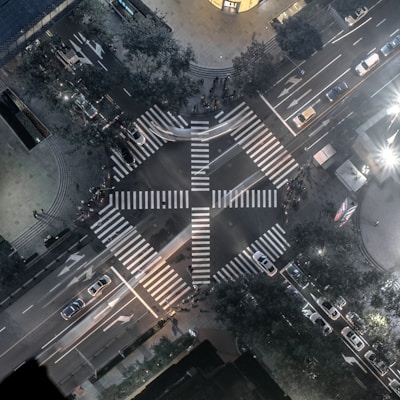 An aerial view of an urban intersection featuring a large, cross-shaped pedestrian crosswalk. The streets are lined with various vehicles, including cars and buses. Trees and buildings border the sidewalks, with some illuminated signage and streetlights visible. The overall scene is bustling yet orderly, with both foot and vehicle traffic.