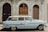 A classic light blue vintage car is parked in front of an ornate, historical building with arched wooden doors and decorative stonework. The car's design features chrome detailing and a roof rack, reflecting mid-20th century style.