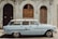 A classic light blue vintage car is parked in front of an ornate, historical building with arched wooden doors and decorative stonework. The car's design features chrome detailing and a roof rack, reflecting mid-20th century style.