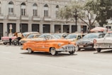 A street scene featuring a lineup of vintage cars parked along the road. In the foreground, a bright orange classic convertible steals the spotlight. The backdrop displays an elegant, historic building with large windows and ornate architectural details. People are seen milling around the cars, some examining an open hood.