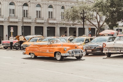 A street scene featuring a lineup of vintage cars parked along the road. In the foreground, a bright orange classic convertible steals the spotlight. The backdrop displays an elegant, historic building with large windows and ornate architectural details. People are seen milling around the cars, some examining an open hood.