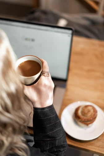 A person enjoying a cup of coffee while browsing a laptop in a bright kitchen.