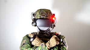 A person wearing camouflage military gear and VR headset, with crossed arms holding controllers. The helmet is equipped with a headset and a glowing red light.