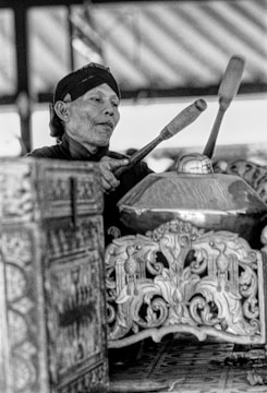 An individual is playing a traditional percussion instrument, known as a gamelan, using mallets. The image captures the focus and concentration of the player, with intricate and ornate carvings visible on the instrument.