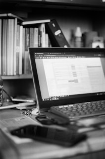 A black and white image of a workspace featuring an open laptop on a desk. The screen displays a spreadsheet or document editor. Surrounding the laptop are various office items such as a smartphone, keys, and a pen. A bookshelf in the background is filled with numerous books and a small spray bottle is visible next to it.