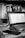 A black and white image of a workspace featuring an open laptop on a desk. The screen displays a spreadsheet or document editor. Surrounding the laptop are various office items such as a smartphone, keys, and a pen. A bookshelf in the background is filled with numerous books and a small spray bottle is visible next to it.
