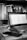 A black and white image of a workspace featuring an open laptop on a desk. The screen displays a spreadsheet or document editor. Surrounding the laptop are various office items such as a smartphone, keys, and a pen. A bookshelf in the background is filled with numerous books and a small spray bottle is visible next to it.
