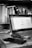 A black and white image of a workspace featuring an open laptop on a desk. The screen displays a spreadsheet or document editor. Surrounding the laptop are various office items such as a smartphone, keys, and a pen. A bookshelf in the background is filled with numerous books and a small spray bottle is visible next to it.