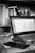 A black and white image of a workspace featuring an open laptop on a desk. The screen displays a spreadsheet or document editor. Surrounding the laptop are various office items such as a smartphone, keys, and a pen. A bookshelf in the background is filled with numerous books and a small spray bottle is visible next to it.